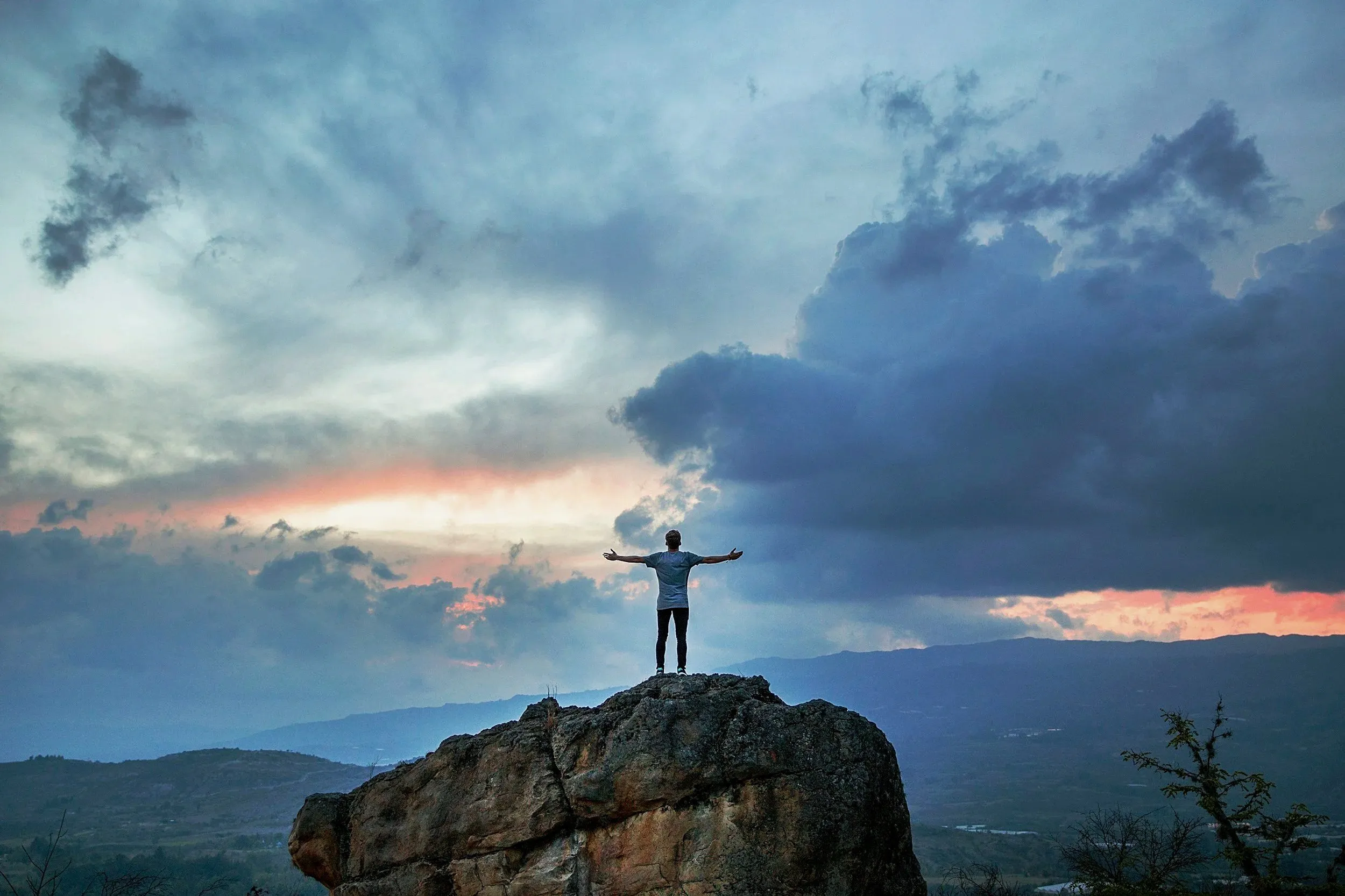 A man with arms spread standing on a mountain precipice at dusk